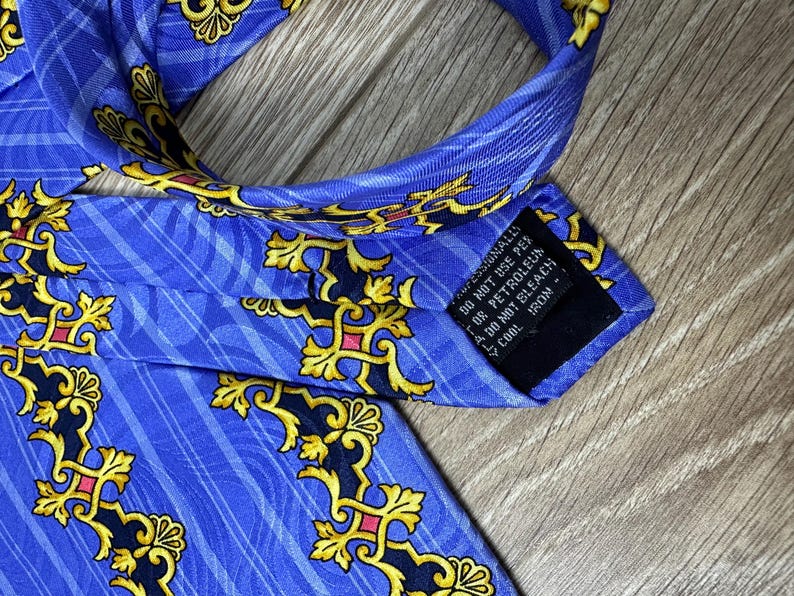 以下が含まれることがあります： 金色と黒の模様が施された青いシルクネクタイ。ネクタイには、「ドライクリーニングのみ、石油系溶剤は使用しないでください、漂白しないでください、低温でアイロンをかけてください。」というケアラベルが付いています。