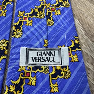 以下が含まれることがあります： 金色と黒の模様が施された青いシルクネクタイ。ネクタイには「Gianni Versace」と書かれたラベルが付いています。