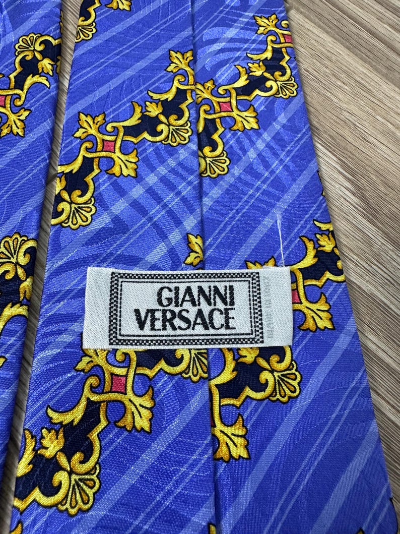 以下が含まれることがあります： 金色と黒の模様が施された青いシルクネクタイ。ネクタイには「Gianni Versace」と書かれたラベルが付いています。
