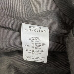 Puede incluir: Primer plano de una etiqueta de prenda de Studio Nicholson. La etiqueta blanca presenta texto negro que detalla la composici&oacute;n de la tela: 60% lana virgen, 40% poliamida y un forro de 50% cupro y 50% algod&oacute;n. Tambi&eacute;n se incluyen instrucciones de cuidado.