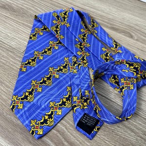 以下が含まれることがあります： 金色と赤色の模様が施された青いシルクネクタイ。ネクタイはストライプの背景に、スタイリッシュな十字架と渦巻きが繰り返されるパターンが施されています。ネクタイには「In Not Just For Any Occasion」と書かれたラベルが付いています。