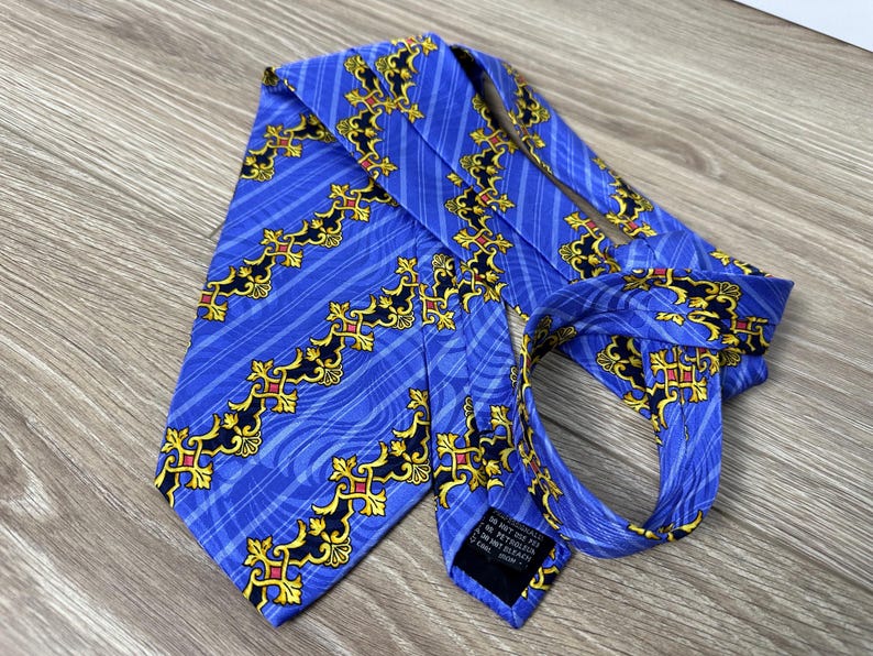 以下が含まれることがあります： 金色と赤色の模様が施された青いシルクネクタイ。ネクタイはストライプの背景に、スタイリッシュな十字架と渦巻きが繰り返されるパターンが施されています。ネクタイには「In Not Just For Any Occasion」と書かれたラベルが付いています。