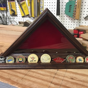 May include: A dark wood shadow box with a red felt lining and glass front. The shadow box is designed to hold and display military challenge coins. The shadow box has eight slots for coins. Seven slots are filled with gold and silver challenge coins. The coins have various designs and text, including "USMC" and "1st Marine Division".