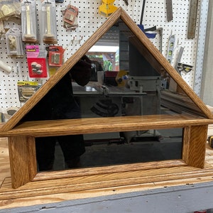 May include: A wooden shadow box with a triangular top and a black glass front. The box has a shelf inside and is made of a light brown wood with a dark stain.
