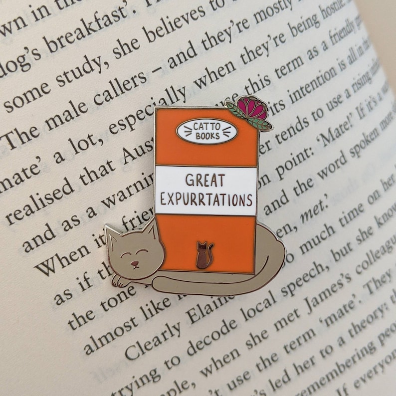 Cat Classics Enamel Pin that reads 'Great Expurrtations', photographed on an open book