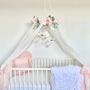 May include: White crib canopy with a floral wreath and a mobile hanging above a white crib with a pink pillow and a white blanket with ruffles.