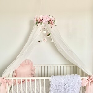 May include: A white canopy with a pink floral garland and a matching floral mobile hangs over a white crib with a pink pillow and a white ruffled blanket.