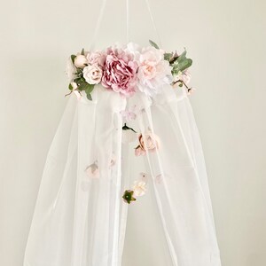 May include: A white canopy with a pink and white floral garland hanging from the top. The garland is made of artificial flowers and greenery.
