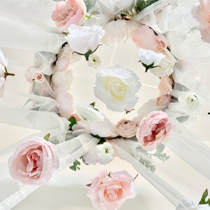 May include: A white tulle canopy decorated with a circle of pink and white flowers. The flowers are arranged in a circular pattern and are interspersed with greenery.