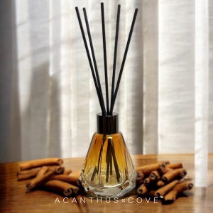 May include: A glass essential oil diffuser with black reeds and a dark metal cap. The amber-colored liquid is visible inside the decorative bottle. Cinnamon sticks are scattered on the wooden surface. The text "ACANTHUS COVE" is printed at the bottom.