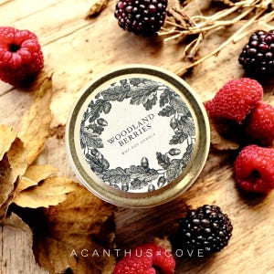 May include: A 6 oz soy candle with a lid featuring the text "Woodland Berries" surrounded by a botanical design. The candle is surrounded by fresh raspberries, blackberries, and autumn leaves on a wooden surface. The brand name "Acanthus Cove" is at the bottom.