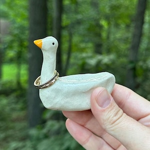 May include: A ceramic goose figurine with a white body, an orange beak, and a gold ring around its neck. The goose is held in a hand against a blurred green background.