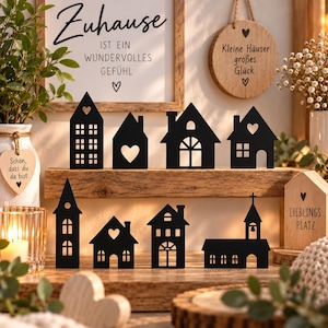 Könnte beinhalten: Eine Sammlung schwarzer Metall-Haus-Silhouetten, die auf zwei Holzregalen angeordnet sind. Die Figuren zeigen ausgeschnittene Fenster, Türen und Herzformen. Dekorative Holzschilder mit deutschem Text und Herzsymbolen sind dazwischen platziert.