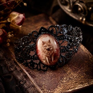 May include: A black metal hair accessory featuring an intricate filigree design and a central oval cabochon. The cabochon displays a detailed illustration of a wolf set against a reddish-brown background, framed by the dark, ornate metalwork.