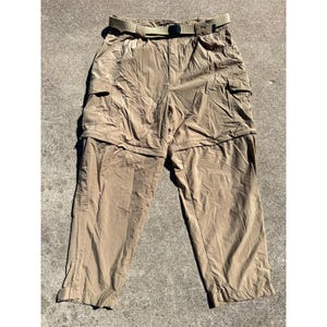 May include: Khaki-colored convertible pants with a matching belt. The pants have multiple pockets and zippers, allowing them to convert into shorts. The belt is a light olive green color and has a black buckle. The pants are made of a lightweight material.