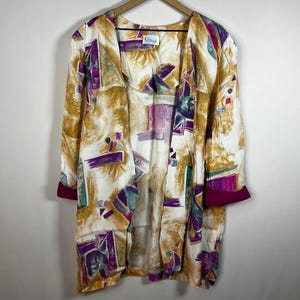 Puede incluir: Una chaqueta vintage abierta con un colorido estampado abstracto. La chaqueta presenta una base color crema con formas geométricas de color mostaza, morado y verde azulado. Los puños son de color magenta intenso. La chaqueta está colgada en una percha de madera.