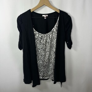 May include: Black short-sleeved cardigan over a scoop-neck top with a silver and gray animal print. The top has a gathered detail at the neckline. The cardigan has a draped front and is hanging on a wooden hanger.