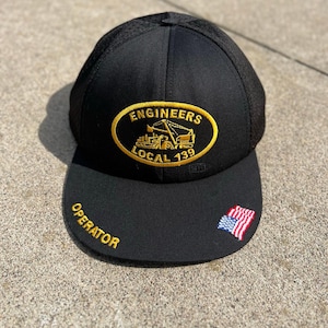 Może przedstawiać: Czarna czapka z daszkiem z siatkowym tyłem i zakrzywionym daszkiem. Czapka posiada haftowaną owalną naszywkę z napisem "ENGINEERS LOCAL 139" i grafiką sprzętu budowlanego. Słowo "OPERATOR" jest wyhaftowane na daszku, wraz z małą amerykańską flagą.
