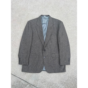 Puede incluir: Un blazer de tweed gris con cierre de dos botones y forro azul claro. El blazer tiene dos bolsillos delanteros y tres botones en cada manga. La chaqueta está tendida sobre una superficie de hormigón.