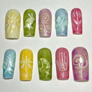 May include: Ten colorful fake nails with different designs. The designs include a sun, a rabbit, a spiral, a butterfly, a flower, a dragonfly, a frog, a handprint, and a snail.