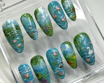 May include: A set of ten almond-shaped press-on nails featuring hand-painted impressionist-style scenes of water lilies, lush green foliage, and a white swan on blue water, all displayed in a clear plastic storage case with a hinged lid.