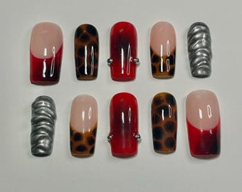 May include: A set of ten press-on nails featuring varied designs including red French tips, brown and black leopard print patterns, solid red nails with small silver studs on the sides, and textured metallic silver nails with a wavy relief.