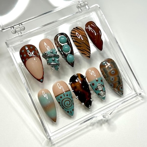 May include: A set of ten artificial nails in a clear plastic case. The nails feature various designs, including teal, brown, and tan colours, with decorative elements like studs, swirls, and animal prints. The nails are almond-shaped.