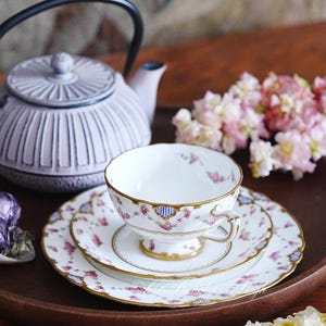 Puede incluir: Un juego de té vintage con una taza, un platillo y un plato blancos, adornados con motivos florales rosas y detalles dorados. También hay una tetera a juego y una bandeja de madera. El conjunto está decorado con flores rosas, creando una estética clásica y elegante.