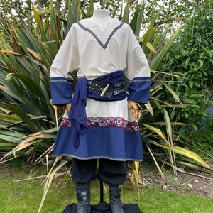May include: A white tunic with blue and red trim, a black leather belt with a silver buckle, and a blue scarf. The tunic is worn over black pants and black boots.