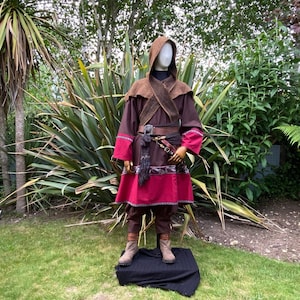 May include: A brown hooded robe with a red and gray trim. The robe has a belt with a silver buckle and a brown scarf. The robe is worn by a mannequin standing on a black platform.