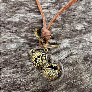 May include: A bronze-colored pendant necklace featuring a detailed, ornate gourd-shaped charm with a fish design. The pendant hangs from a brown leather cord. The necklace is displayed on a textured, gray and brown surface.