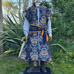 Puede incluir: Un abrigo largo azul con un patrón de damasco dorado, usado sobre una camisa de manga larga gris claro. El conjunto incluye un cinturón de cuero negro con hebilla dorada, superpuesto con una faja de tela naranja y azul. Un colgante cuelga al cuello.