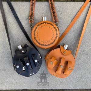 May include: Three leather flasks with shoulder straps. One is black, and two are tan. The tan flasks have decorative stitching and small pouches. The flasks have silver metal tops and are round in shape.