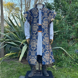 May include: A long, ornate blue and gold patterned coat with a high collar and wide sleeves. The coat is worn over a light gray shirt and black pants. The outfit is displayed on a mannequin outdoors.