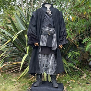 May include: A black costume ensemble featuring a long coat with velvet cuffs, a patterned vest, and matching trousers. The outfit includes a wide belt with a silver buckle and black boots. The costume is displayed on a mannequin.