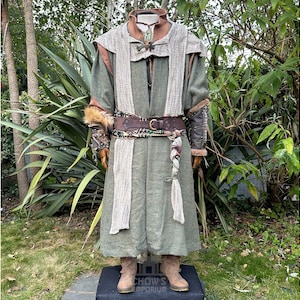 May include: A long olive green tunic with a textured beige shawl draped over the shoulders. The outfit includes a brown leather belt with a metal buckle, dark brown arm guards with fur trim, and tan boots. A patterned sash hangs from the waist.