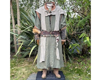 May include: A long olive green tunic with a textured beige shawl draped over the shoulders. The outfit includes a brown leather belt with a metal buckle, dark brown arm guards with fur trim, and tan boots. A patterned sash hangs from the waist.