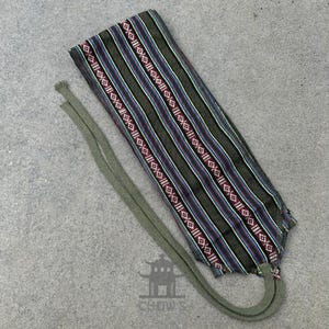 May include: A green, purple and red striped fabric headband with a long green tie. The headband is made of woven fabric and has a geometric pattern.