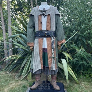 May include: A layered outfit featuring a long olive green coat with tan cuffs, a textured grey hooded shawl, a tan tunic, and dark striped trousers. A black leather belt with a gold buckle cinches the waist. Brown boots complete the ensemble.