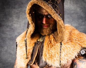 May include: A dark brown hooded cowl featuring a thick tan faux fur trim along the hood and shoulder cape. The hood includes decorative gold-toned metal studs and brown cord lacing details. Two long, thin braided cords with small metal charms hang down.