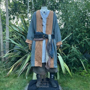Pode incluir: Um traje de estilo medieval exibido em um manequim. O traje apresenta um casaco longo cinza e marrom com botões de madeira, um cinto preto e um lenço azul. O manequim está em uma plataforma preta com o texto "CHOW'S EMPORIUM".