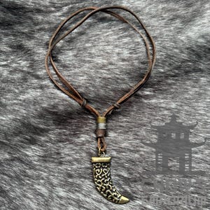 May include: A brown leather cord necklace with a decorative metal pendant shaped like a horn. The pendant has an intricate design and a gold finish.