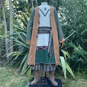 Puede incluir: Un disfraz de estilo medieval exhibido en un maniquí. El atuendo incluye un abrigo largo verde con ribetes marrones, una túnica blanca con cuello en V, un cinturón estampado y pantalones a rayas. Botas marrones completan el conjunto. Un collar cuelga del maniquí.