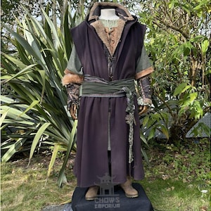 Pode incluir: Uma longa túnica de cor roxa escura com uma faixa verde e detalhes em couro marrom. A túnica tem um capuz e é usada sobre um colete de couro marrom. A pessoa está em pé em frente a um fundo verde frondoso.