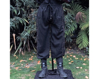 May include: Black trousers featuring an elasticated waistband with a drawstring closure and decorative side panels with vertical lace-up detailing. The trousers have a straight-leg cut and are paired with black boots featuring two horizontal straps.