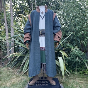 May include: A teal-colored, long-sleeved robe with brown accents on the shoulders and sleeves. The robe is worn over a white tunic with a V-neck and a decorative belt. A necklace with a pendant is visible. The outfit is displayed on a mannequin.