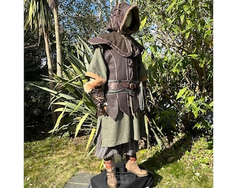 May include: A hooded, brown leather vest with fur trim, a green tunic, and brown leather pants. The vest has a belt with a buckle and a decorative design. The tunic has a long, flowing skirt and a fur-trimmed collar. The pants are cinched at the waist with a belt.