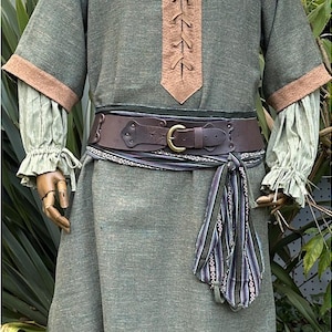 Puede incluir: Una túnica de estilo medieval en verde oliva con ribetes marrones y un escote con cordones. El atuendo incluye un cinturón de cuero marrón con hebilla de latón y una faja estampada. Las mangas son de color verde claro con puños con volantes.