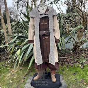 May include: A medieval-style costume displayed on a mannequin. The outfit includes a long brown tunic, a tan coat with a patterned trim, and brown pants. A brown belt with a gold buckle cinches the waist. The costume is completed with brown boots.