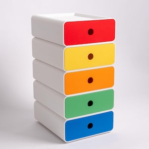 Può includere: Una pila di cinque cassetti bianchi con frontali colorati. I frontali dei cassetti sono rossi, gialli, arancioni, verdi e blu, ciascuno con una maniglia circolare nera. I cassetti sono impilati verticalmente su uno sfondo bianco.